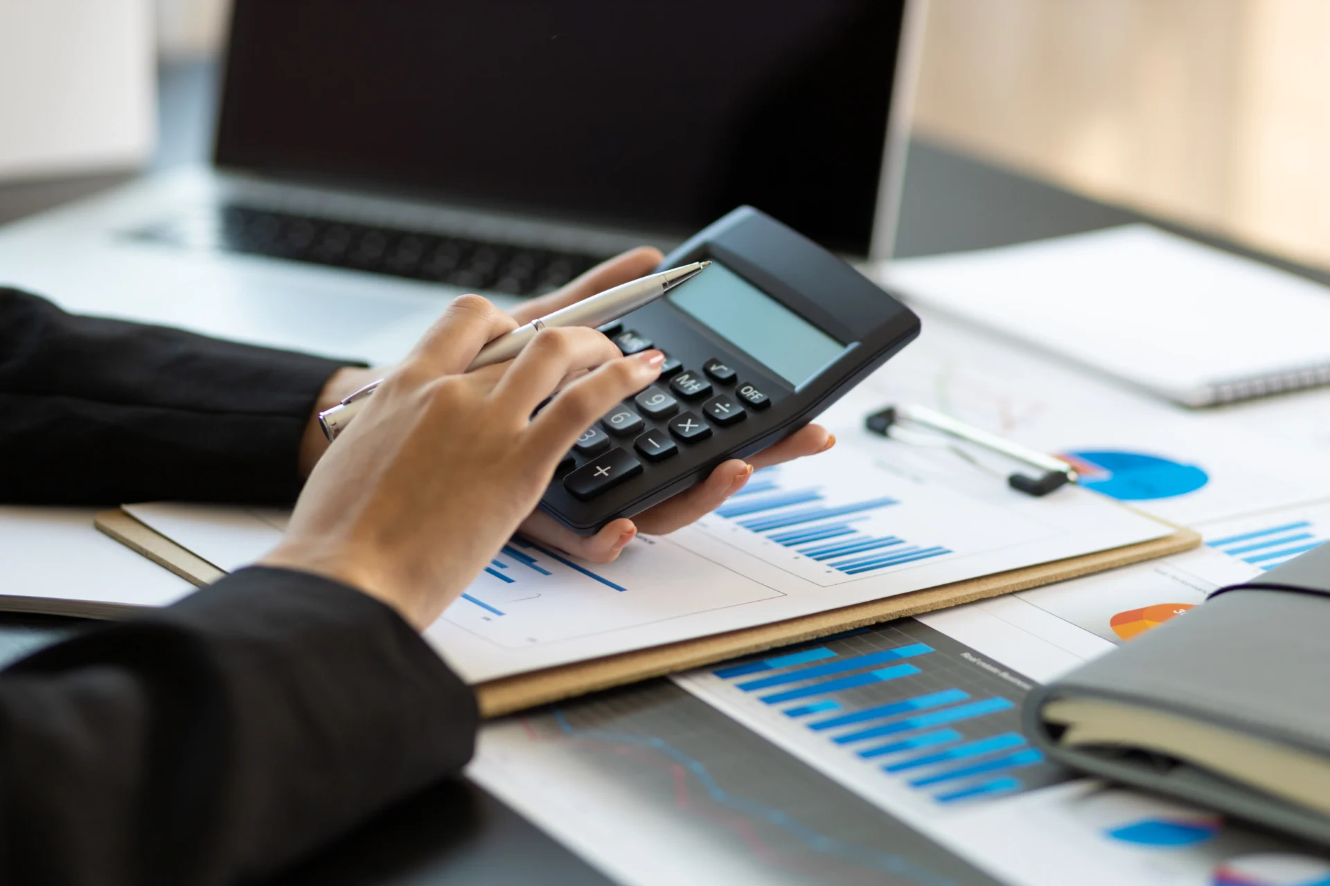 1742465866089_IMAGE-51614++-++ProfitMax+Profes Person using a calculator with charts, graphs, and a laptop on the desk.