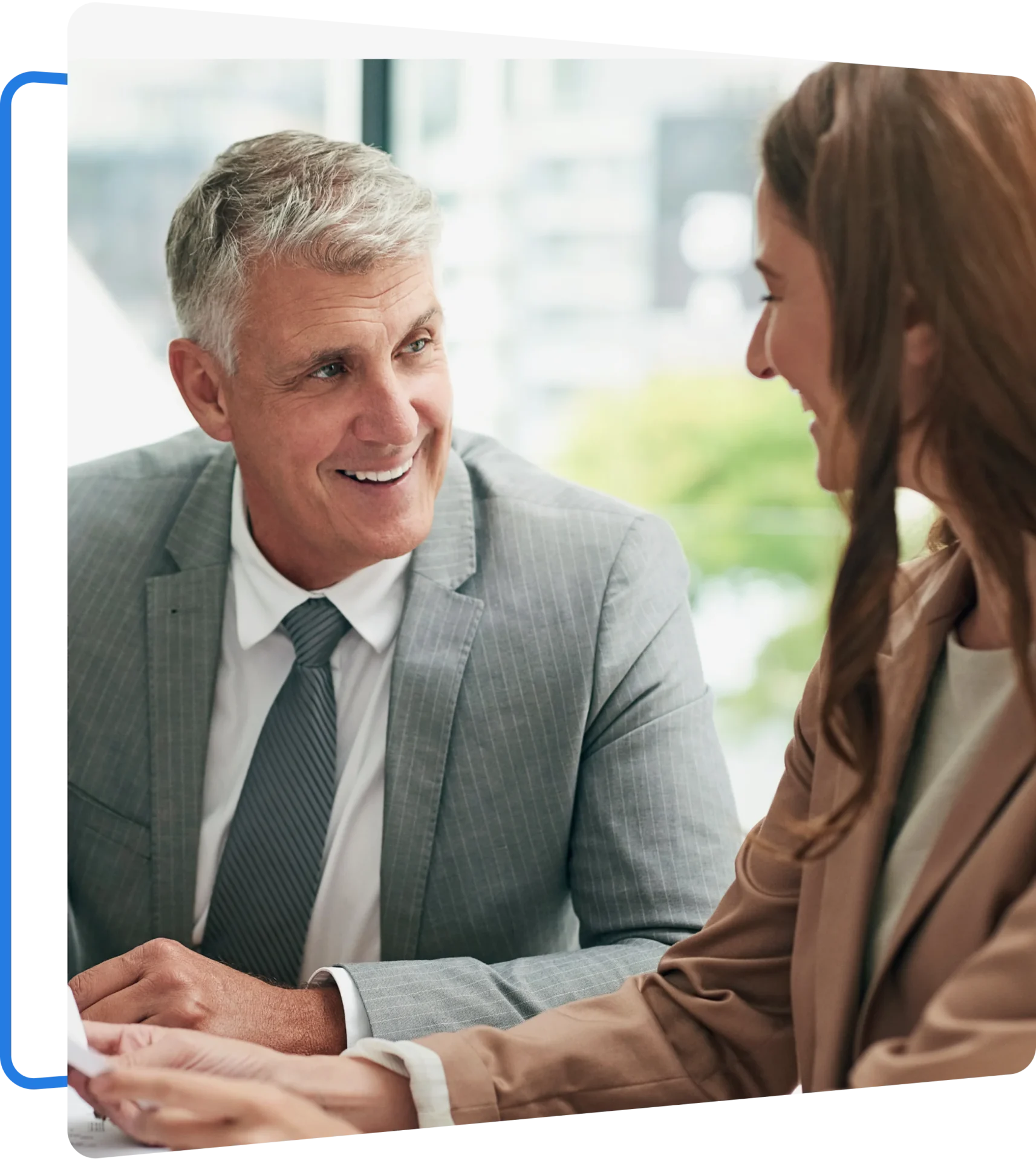 Group 5 (2) A man and a woman in business attire are sitting and smiling at each other while engaging in conversation in an office setting.