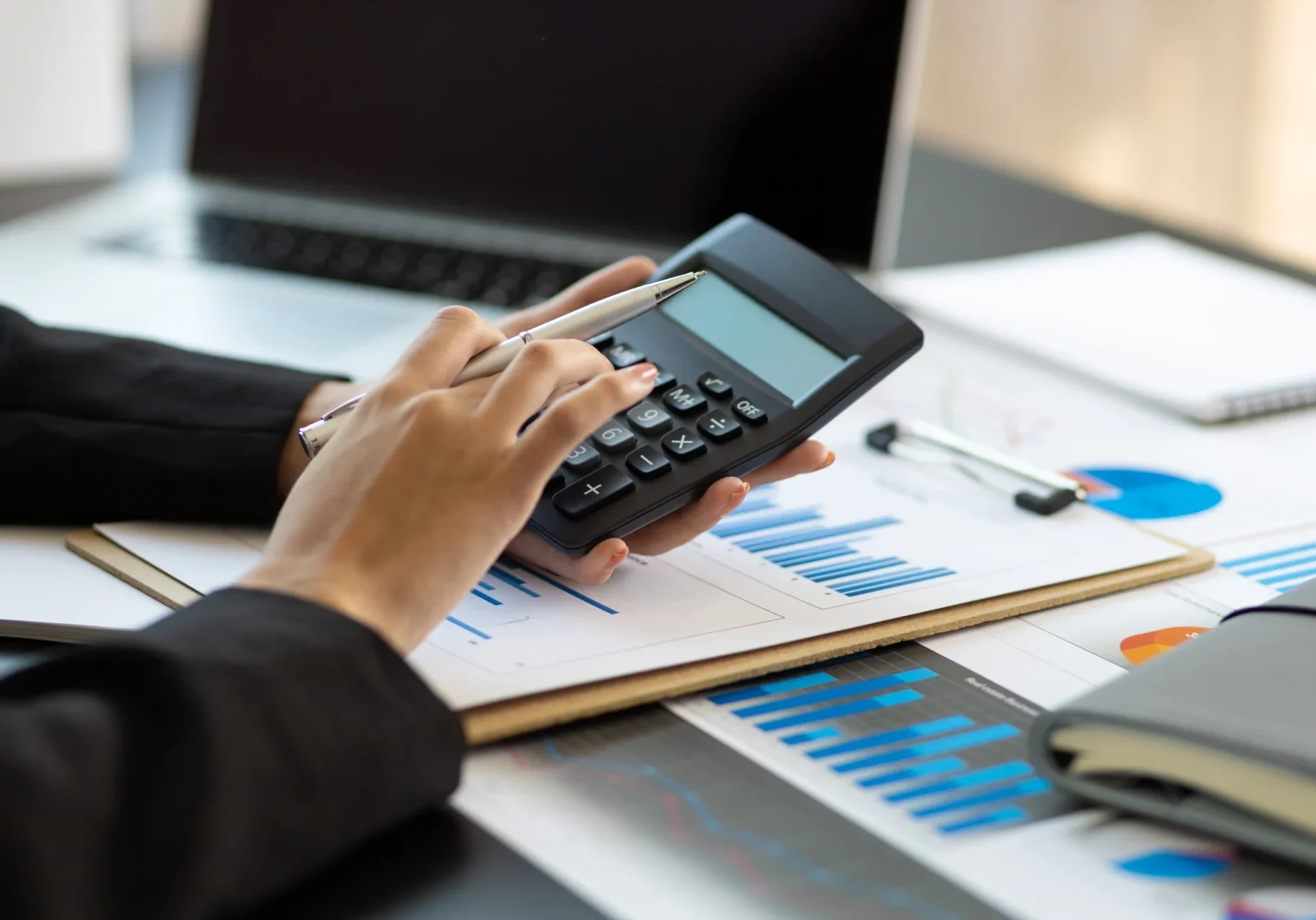 1742465866089_IMAGE-51614++-++ProfitMax+Profes Person using a calculator with charts, graphs, and a laptop on the desk.